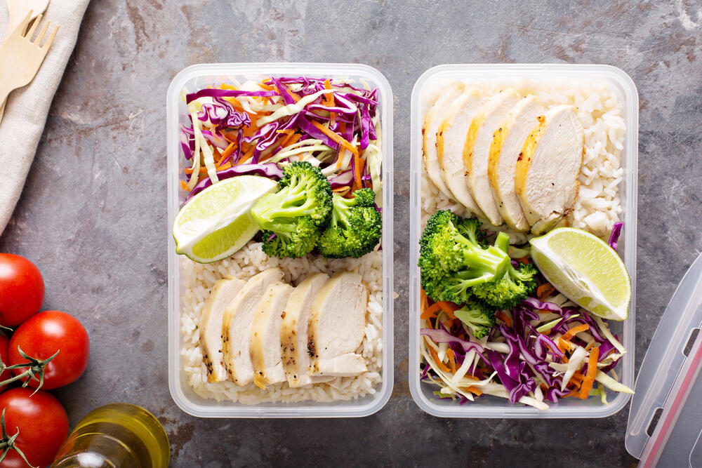Two neatly packed meal prep containers filled with a balanced and nutritious meal sit on a textured grey surface. Each container contains sliced grilled chicken breast on a bed of white rice, vibrant steamed broccoli, and a fresh slaw made of shredded red and white cabbage with thinly sliced carrots. A lime wedge is placed on top for added zest and flavor. Surrounding the containers are whole cherry tomatoes on the vine, a bottle of olive oil, and wooden cutlery, reinforcing a healthy, homemade approach to meal planning. This visually appealing meal prep highlights the importance of portion control and nutrient-dense foods for effective weight loss and maintaining a healthy diet