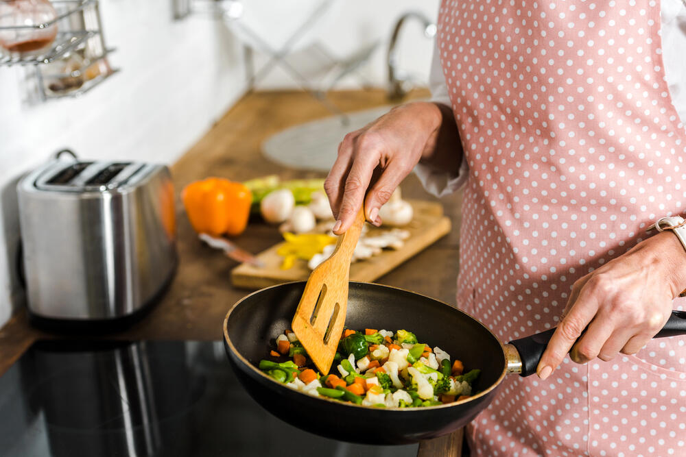 A close-up of a woman cooking a nutritious meal in a modern kitchen. She is wearing a pink polka-dot apron and stirring a pan filled with colorful vegetables, including chopped carrots, broccoli, onions, and green peppers, using a wooden spatula. The stovetop is clean and sleek, reflecting a mindful approach to healthy cooking. In the background, a wooden cutting board holds fresh ingredients such as yellow and orange bell peppers, mushrooms, and a knife, reinforcing the emphasis on fresh, whole foods. A stainless steel toaster sits on the countertop, adding to the cozy, homey atmosphere. This image perfectly illustrates the benefits of preparing home-cooked, nutrient-dense meals as part of a weight loss and healthy lifestyle routine