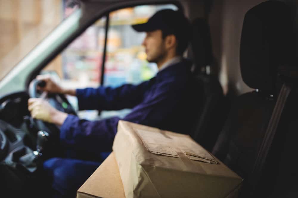 Delivery driver driving van with parcels on seat outside warehouse