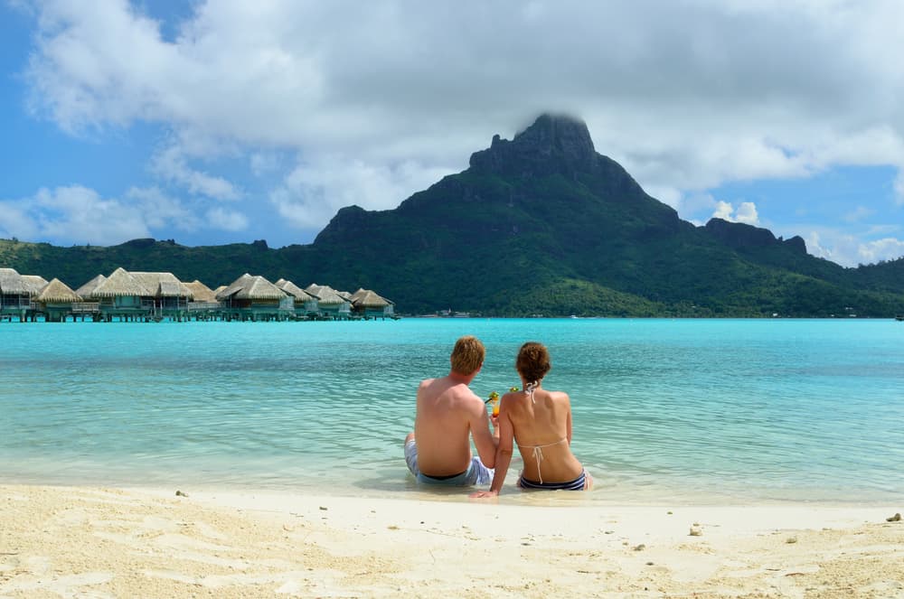A honeymoon couple drinking a cocktail on the beach of a luxury vacation resort in the lagoon with a view on the tropical island of Bora Bora, near Tahiti, in French Polynesia.
