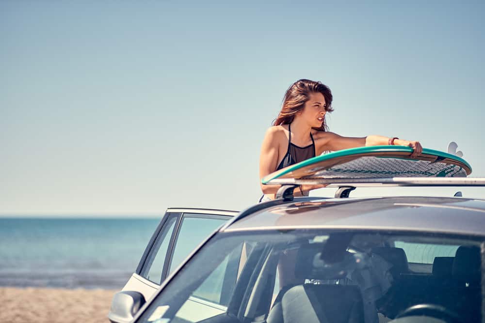 Summer holiday road trip vacation- Young smiling surfer girl at the beach with her surfboard on car Vacation. Extreme Sport