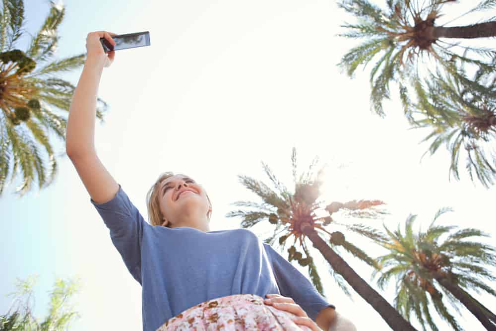 Under view of a young tourist woman raising her arm and holding and pointing a smartphone device to take pictures of palm trees against a sunny blue sky on a summer holiday, outdoors. Travel and technology lifestyle.