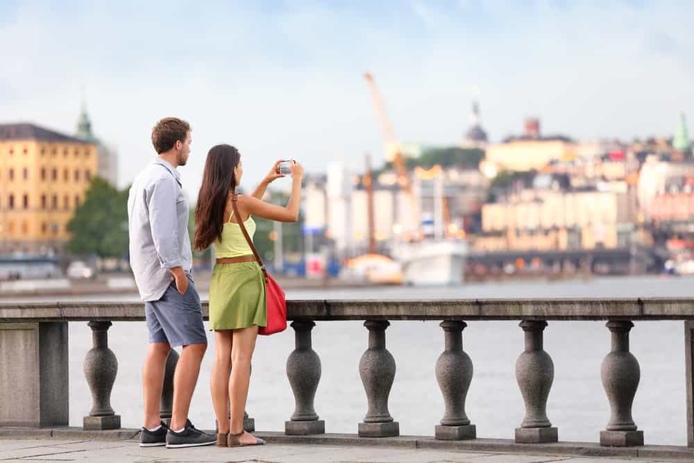 Europe travel tourist people taking pictures. Tourists couple in Stockholm taking smartphone photos having fun enjoying skyline view and river by Stockholm's City Hall, Sweden.