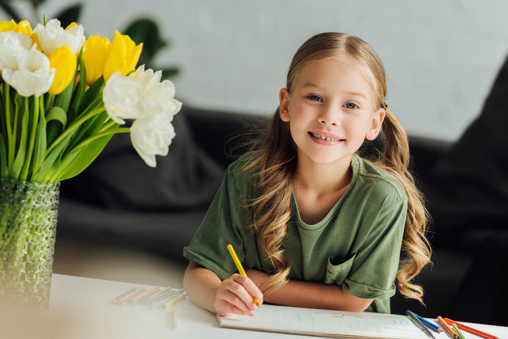 Smiling young girl with long blonde hair drawing with colored pencils at a table, next to a vase of fresh yellow and white tulips, creating a bright and cheerful home learning or creative activity scene