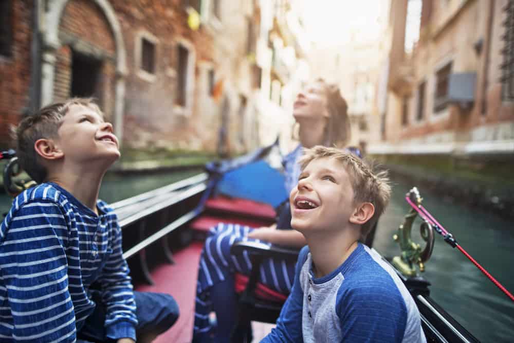 Kids visiting Venice, Italy. They are looking up and smiling in gondola boat during a ride through city canals.