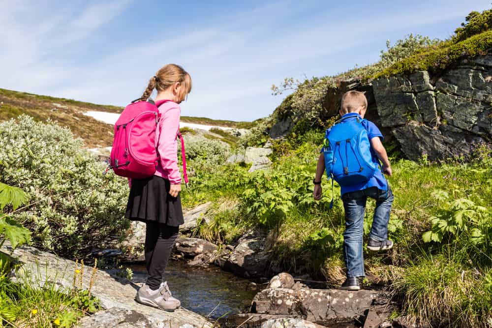 Two kids on a hike wearing their backpacks and exploring nature in Norway on a sunny day with remains of snow in the background.