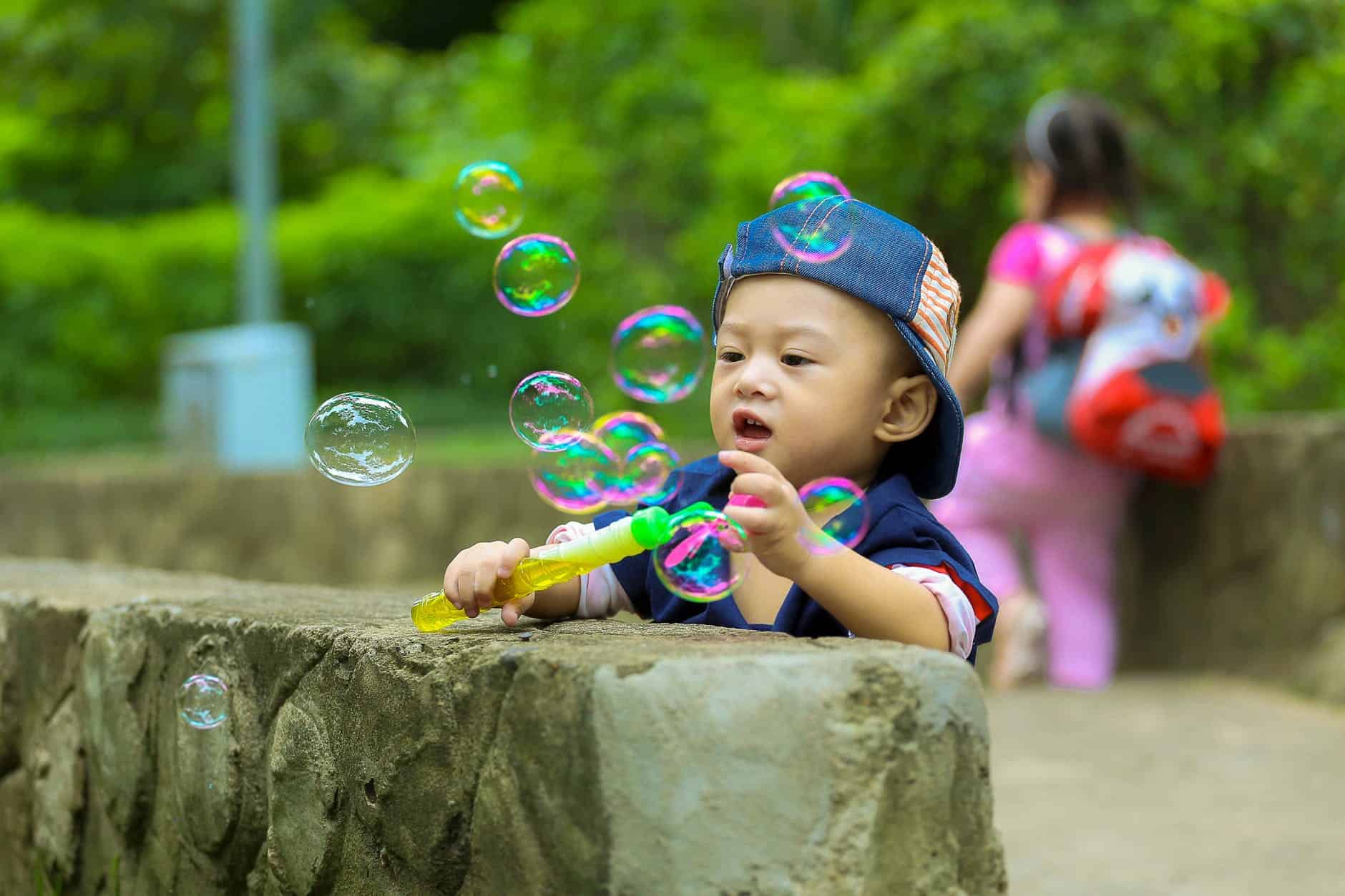 boy in blue fitted cap playing bubbles and leaning on grey concrete wall at daytime