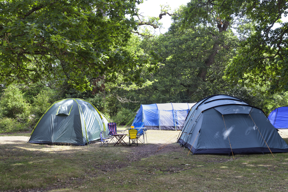 Blue and green family camping tents with table and chairs, under oak trees in the woodland campground, on a sunny summer day .