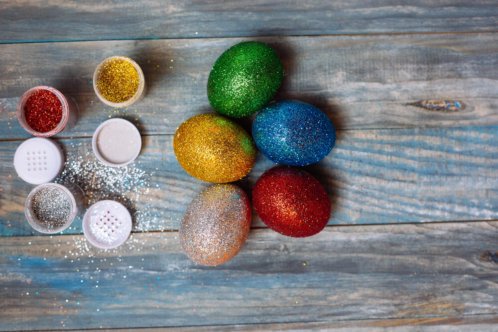 A creative Easter egg decorating setup featuring six glitter-covered eggs in vibrant colors—green, blue, red, gold, and silver—arranged on a rustic wooden surface. Surrounding the eggs are small open containers of fine glitter in shades of red, gold, silver, and white, with some glitter spilled onto the table. The scene showcases a DIY craft project, highlighting a fun and festive way to decorate Easter eggs with a sparkling touch. The wooden background adds a warm, rustic contrast to the bright, shimmering decorations.