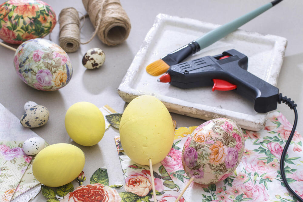 A DIY Easter egg decorating setup featuring the decoupage technique. Several eggs are in different stages of decoration—some painted solid yellow, while others are covered with floral-patterned napkins. The workspace includes essential crafting tools such as a hot glue gun, a paintbrush, and rolls of natural twine. Delicate floral napkins with pink, yellow, and green hues are scattered around, ready to be used for the decoupage process. Small speckled quail eggs add a natural touch to the arrangement. The scene captures a creative and artistic approach to Easter egg decoration, blending rustic and elegant aesthetics.