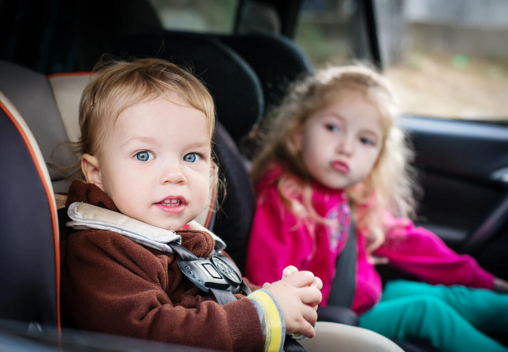 A close-up of two young children securely seated in the back of a used family car, highlighting the importance of safety when choosing a pre-owned vehicle. The toddler in the foreground, with bright blue eyes and light brown hair, is strapped into a well-padded car seat with a five-point harness, wearing a brown fleece jacket. He gazes curiously at the camera with a slight smile. Behind him, an older sibling with curly blonde hair sits in a booster seat, dressed in a vibrant pink jacket and green pants. She wears a seatbelt and appears slightly out of focus, looking off to the side. The car's interior is clean and spacious, with proper child safety seats in place. Through the car window, a blurred outdoor setting is visible. This image reinforces the importance of ensuring a used family car has sufficient space for child seats and meets essential safety standards to protect young passengers on every journey