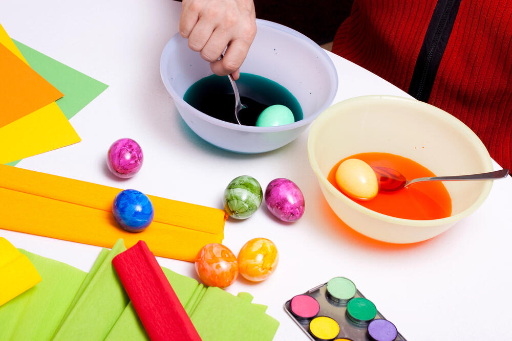 A close-up photograph of a person dyeing Easter eggs using bowls of colored water. The person, wearing a red sweater, is dipping a light green egg into a bowl of deep blue dye with a spoon, while another egg sits in a separate bowl filled with bright orange dye. Several marbled Easter eggs in shades of pink, green, blue, orange, and yellow are placed on the white table. Surrounding the eggs are craft materials, including sheets of colorful crepe paper in red, yellow, green, and orange, along with a watercolor paint palette. The setup suggests a fun and creative Easter egg decorating session.