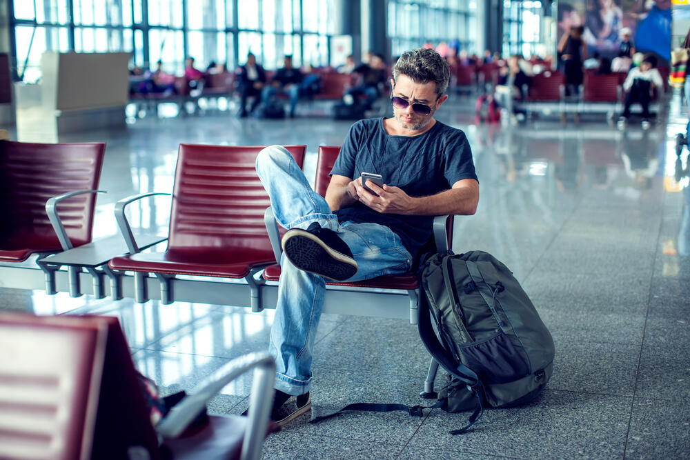A young man with light skin, short, slightly messy dark hair, and a casual appearance is sitting in an airport waiting area. He is dressed in a dark gray t-shirt, slightly faded blue jeans, and black sneakers with white soles. He wears sunglasses with dark lenses and a thin metallic frame, and he is leaning back in a red cushioned chair with metal armrests, one leg crossed over the other in a relaxed posture. He holds a smartphone in both hands, appearing focused on the screen. Beside him, a large greenish-gray backpack with black straps and multiple compartments is resting on the chair. A strap dangles onto the floor, indicating that it is loosely placed rather than fully secured. The background shows a busy airport terminal with other travelers sitting or walking around, large windows allowing natural light to stream in, and a polished floor reflecting the overhead lights. The atmosphere suggests a bustling yet calm setting, with passengers waiting for their flights.