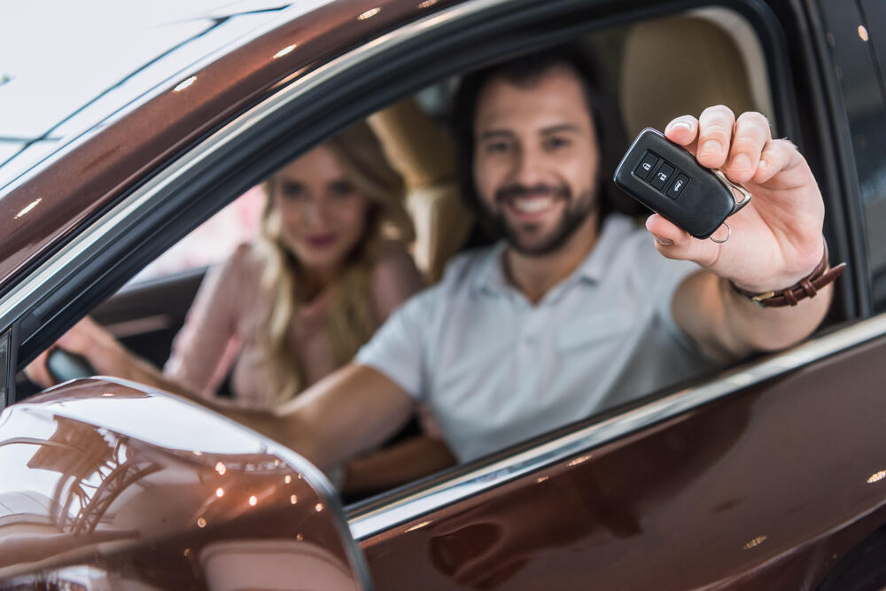 A smiling couple sits inside a recently purchased used car, celebrating their new vehicle choice. The man in the driver’s seat, wearing a white polo shirt and a wristwatch, excitedly holds up the car key fob towards the camera, showcasing their successful purchase. His partner, a blonde woman dressed in a soft pink blouse, sits beside him in the passenger seat, slightly blurred but visibly pleased. The interior of the car appears clean and well-maintained, with tan leather seats, reflecting their careful selection process. The setting is indoors, possibly a dealership or showroom, with bright lighting highlighting the car’s glossy brown exterior. This image captures the excitement of buying a reliable used family car, reinforcing the importance of making an informed decision and taking the time to find the perfect vehicle