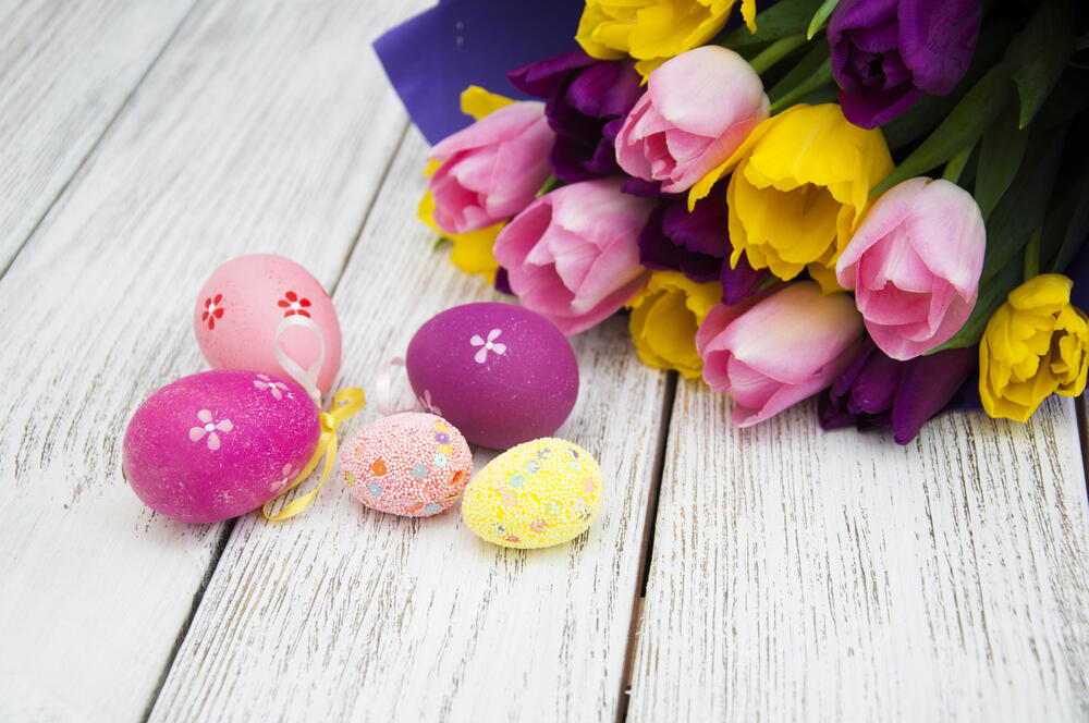 A beautifully arranged Easter-themed composition featuring decorated eggs and a bouquet of tulips on a rustic white wooden surface. The eggs are in various shades of pink and purple, adorned with delicate white floral patterns and tiny decorative beads. Some eggs have small ribbons attached, suggesting they may be used as hanging ornaments. Beside the eggs, there is a vibrant bouquet of tulips in shades of pink, yellow, and deep purple, partially wrapped in blue paper. The combination of bright flowers and creatively decorated eggs evokes a festive and springtime atmosphere, perfect for Easter celebrations.