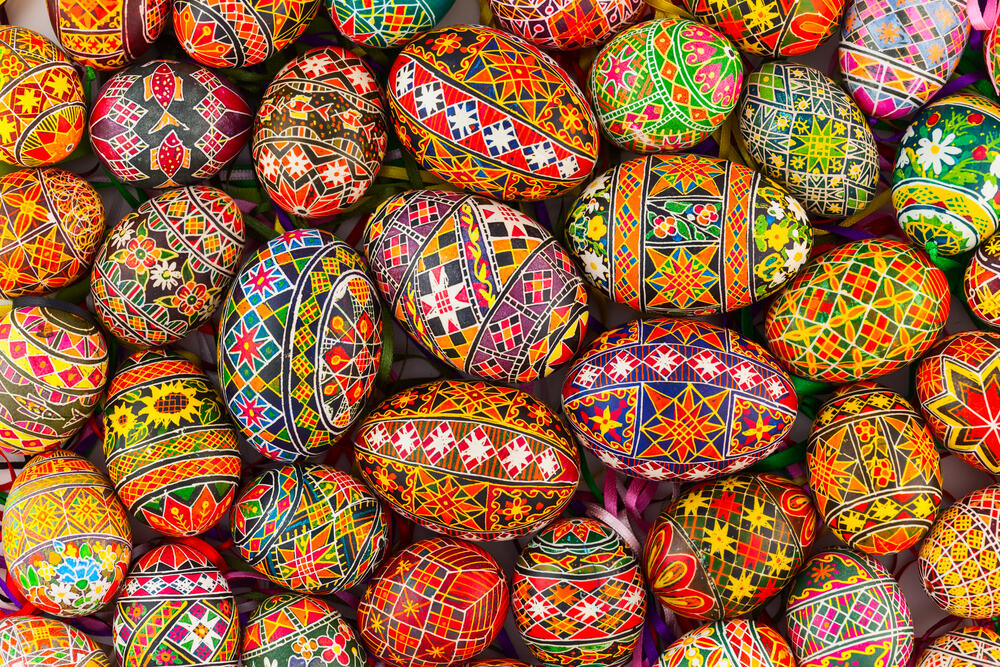 A vibrant collection of hand-decorated Easter eggs featuring intricate and colorful traditional patterns. Each egg is adorned with detailed geometric shapes, floral motifs, and symmetrical designs in shades of red, orange, yellow, blue, green, and black. The eggs showcase the art of Pysanky, a traditional Ukrainian egg-decorating technique that uses wax-resist dyeing to create elaborate and symbolic patterns. Some eggs have delicate ribbon loops attached, suggesting they may be used as hanging ornaments. The overall composition is visually striking, highlighting the craftsmanship and cultural significance of these beautifully decorated eggs.