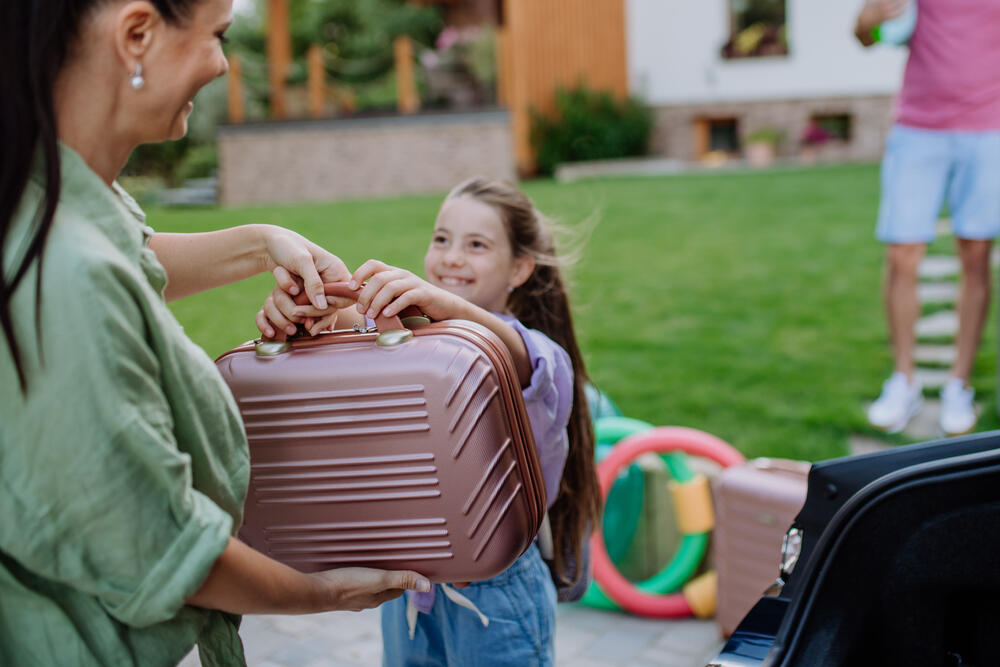 Smiling mother and daughter packing suitcase into car for family holiday, preparing home before travel