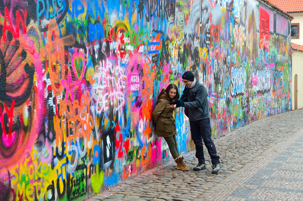 Colorful graffiti-covered Lennon Wall in Prague, Czech Republic, with two tourists admiring street art and messages of peace and freedom