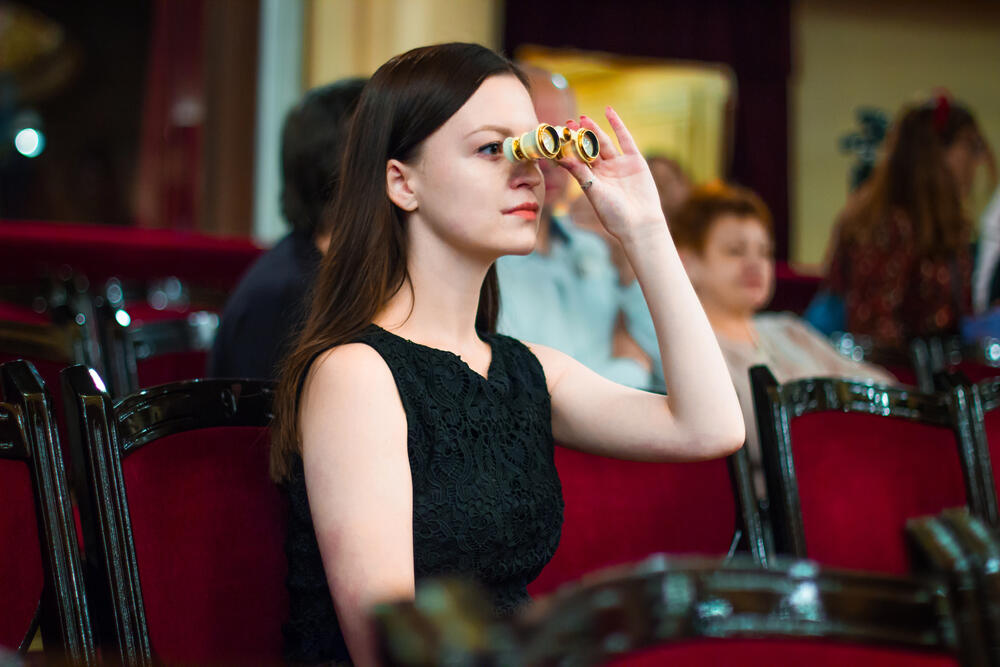 Elegant woman in a black dress using opera glasses while seated in a red velvet theatre, preparing to watch a live stage performance.