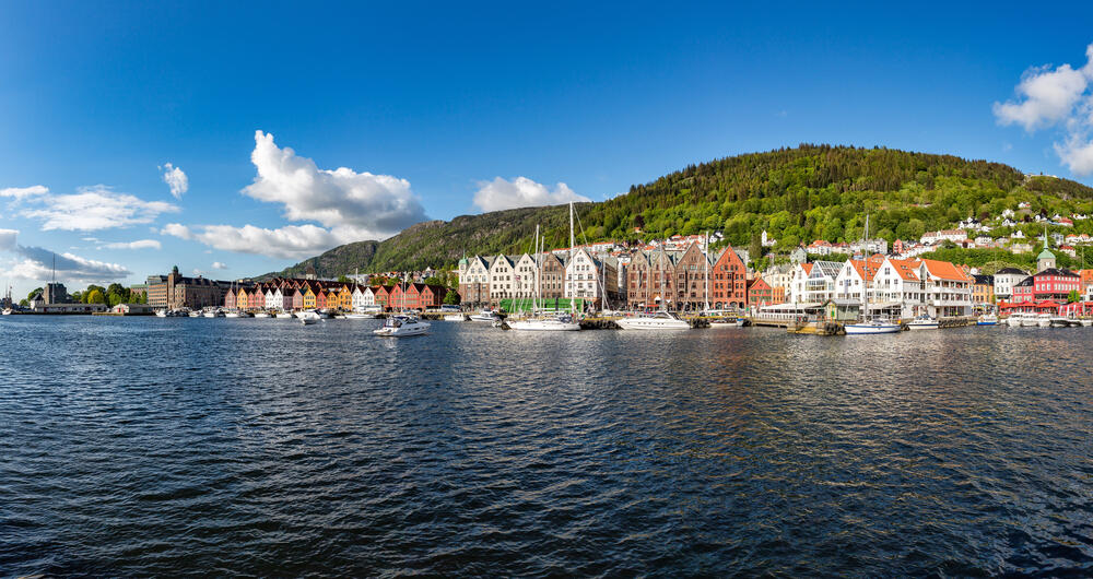 Panoramic view of Bergen’s historic Bryggen waterfront in Norway, featuring colorful wooden buildings, boats in the harbor, and forested hills under a clear blue sky.