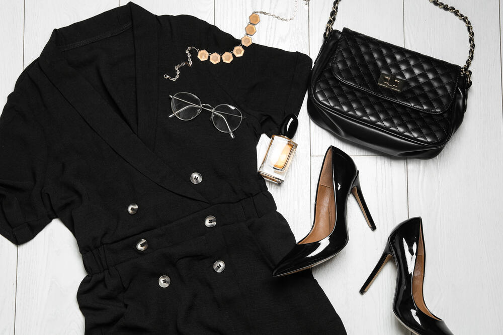 Flat lay of a stylish black theatre outfit featuring a tailored jumpsuit, patent leather high heels, quilted handbag, glasses, perfume, and gold necklace on a white wooden background.