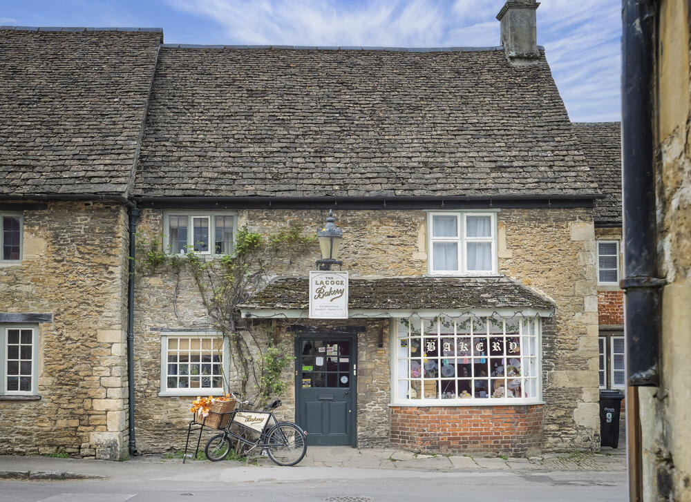 Historic stone bakery storefront in Lacock Village, Cotswolds, England, with vintage bicycle display and traditional window showcasing baked goods.