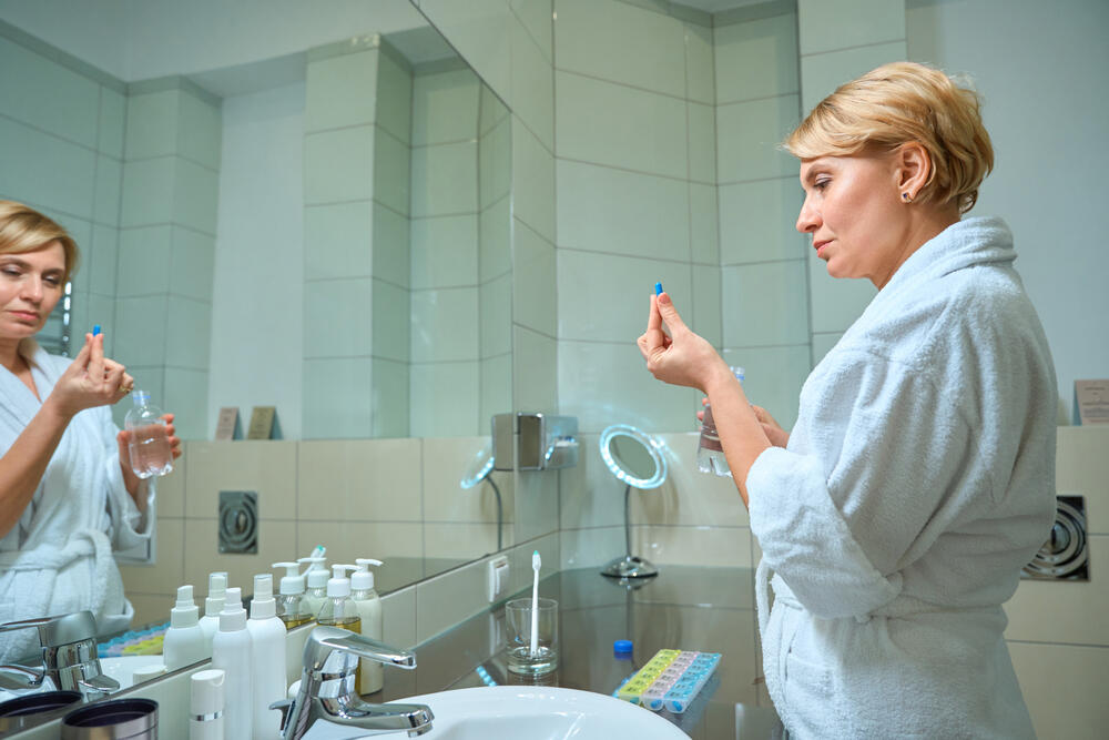 Middle-aged woman in a hotel bathroom holding a supplement pill and glass of water, standing by a travel pill organizer