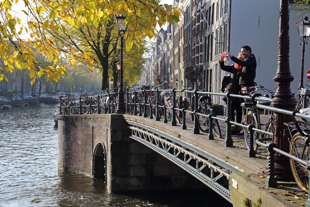 Tourists taking a selfie on a canal bridge in Amsterdam during autumn, with bikes, historic buildings, and golden leaves in the background