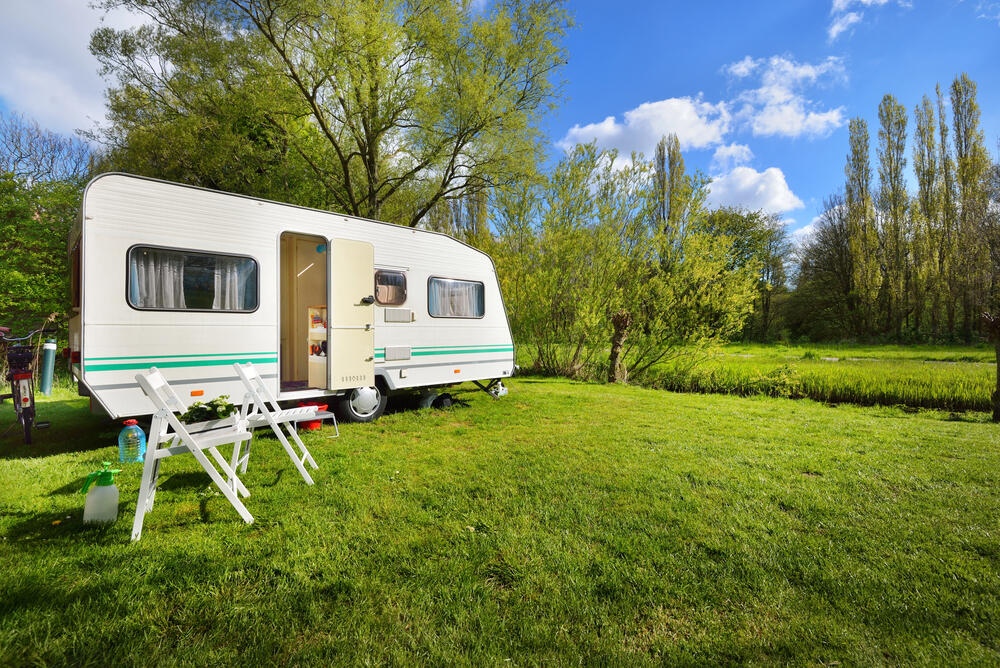 White caravan trailer with open door and folding chairs set on a grassy campsite surrounded by lush trees under a blue sky, ideal for nature-focused travel and eco-friendly camping.