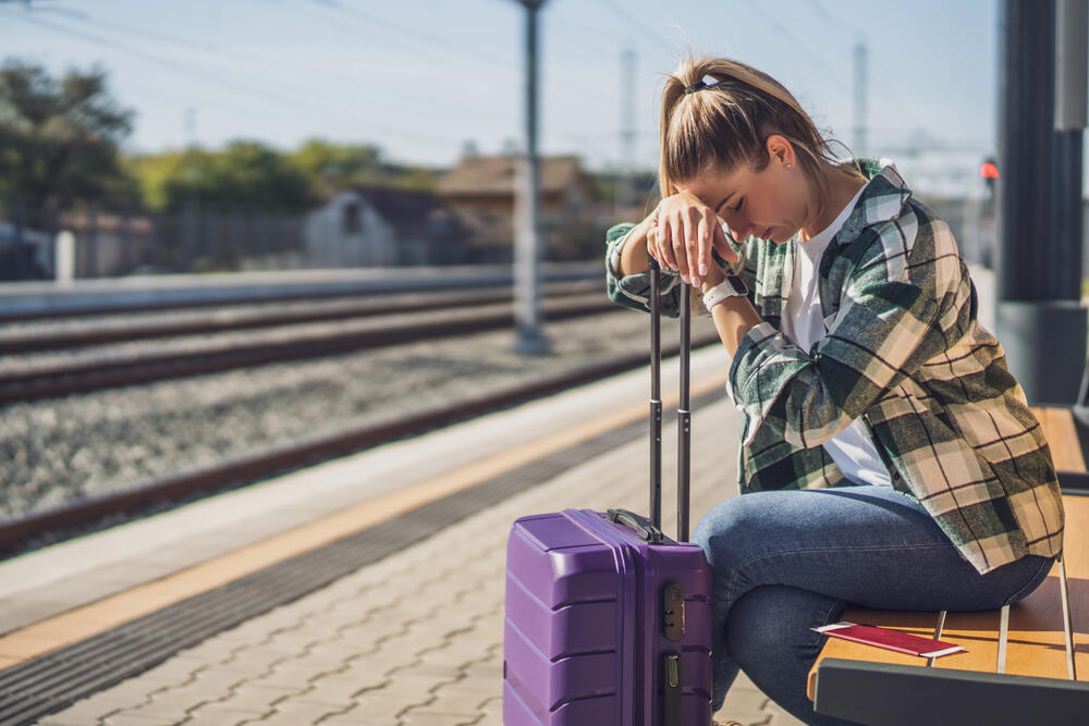 Stressed young woman sitting on a bench at a train station with her travel suitcase, appearing worried or exhausted during her journey and may have lost phone