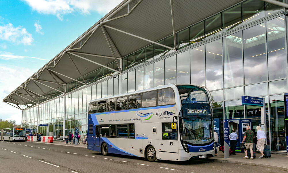Airport Flyer A1 double-decker bus outside the glass-fronted terminal building at Bristol Airport, with passengers boarding and clear signage for the Airport Express Link to Bristol city centre.