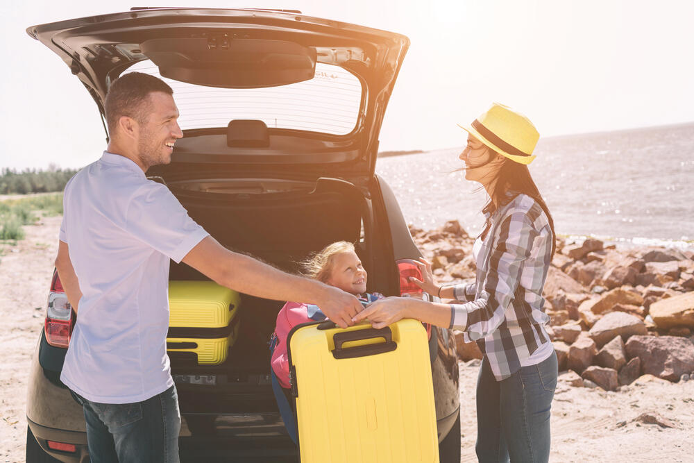 Happy family unloading yellow suitcases from car boot during a summer beach road trip, with parents smiling and child sitting inside the trunk
