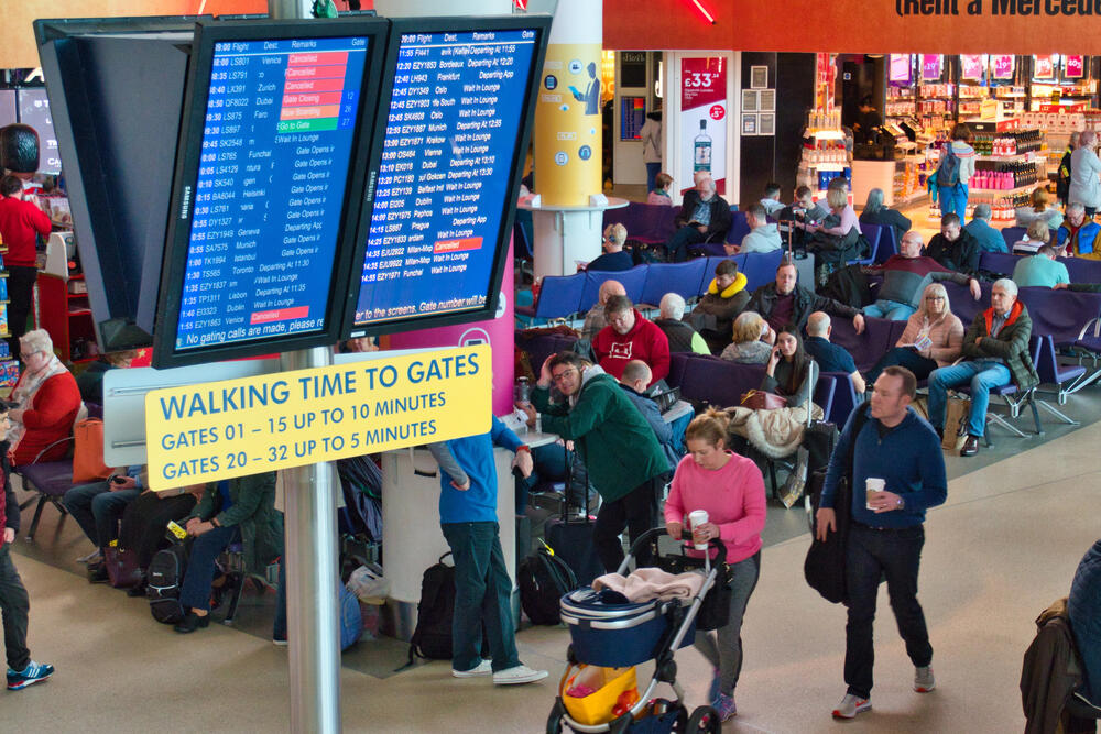 Busy departure lounge at Manchester Airport with digital flight information screens, walking time signage to gates, families and travellers seated or moving with luggage and coffee, capturing the hustle of airport travel.