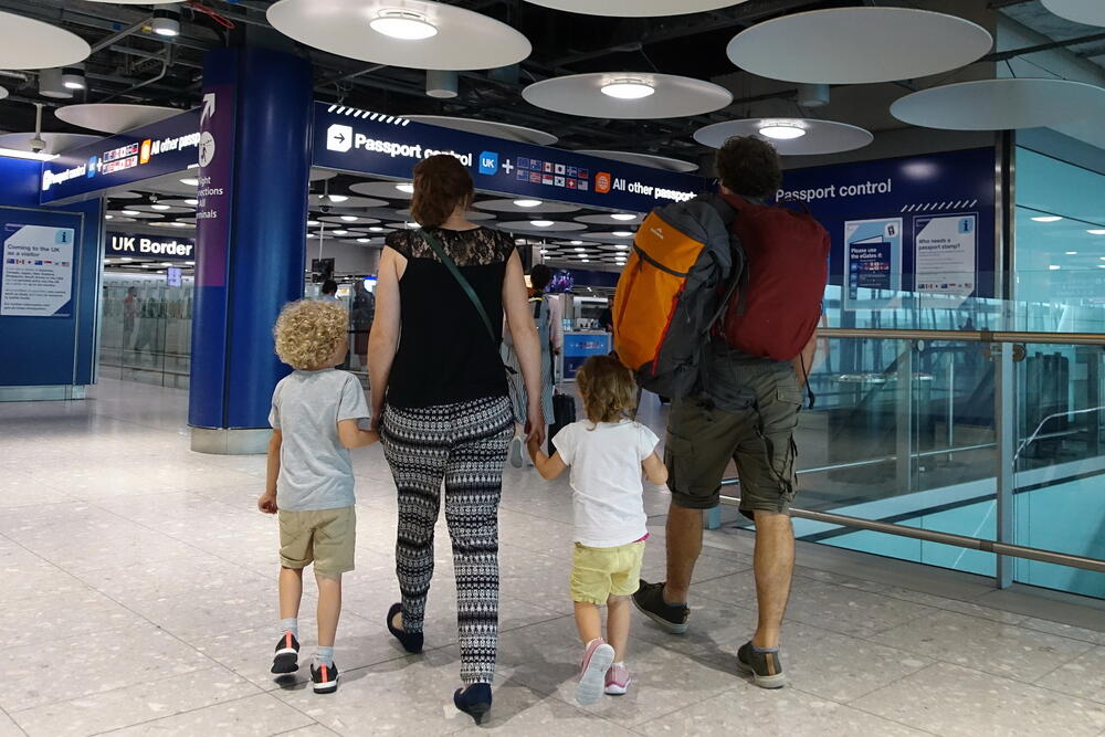 Family with young children walking towards passport control at a UK airport, carrying travel backpacks and hand-in-hand, illustrating a typical family travel experience through border control and arrivals.