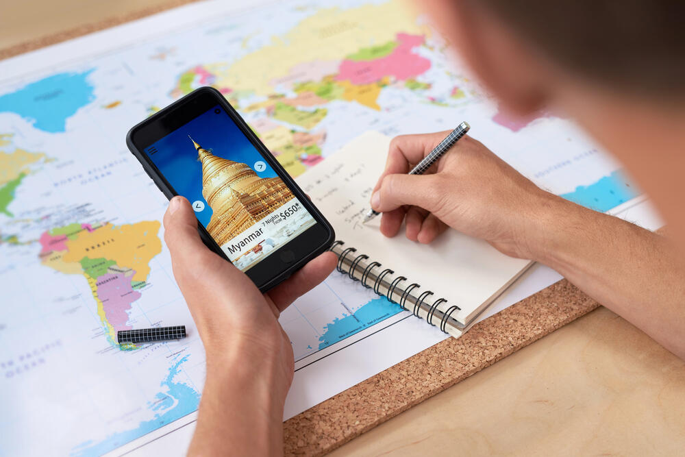 Person planning a trip using travel apps on a smartphone while writing travel notes next to a world map