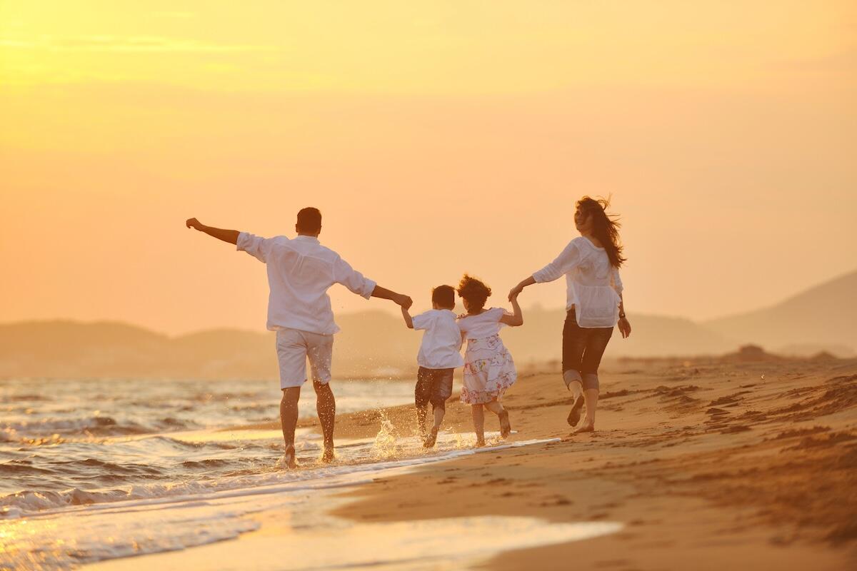 A joyful family of four runs along the beach at sunset, holding hands and splashing in the waves—capturing the essence of a carefree, budget-friendly vacation.