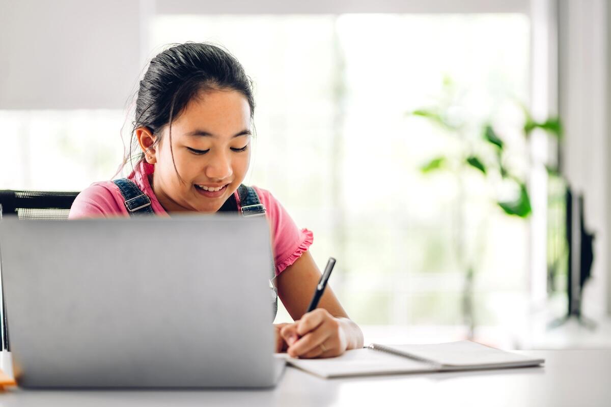 Smiling young girl studying online at home, writing in a notebook while looking at her laptop screen, with natural light and a plant in the background.