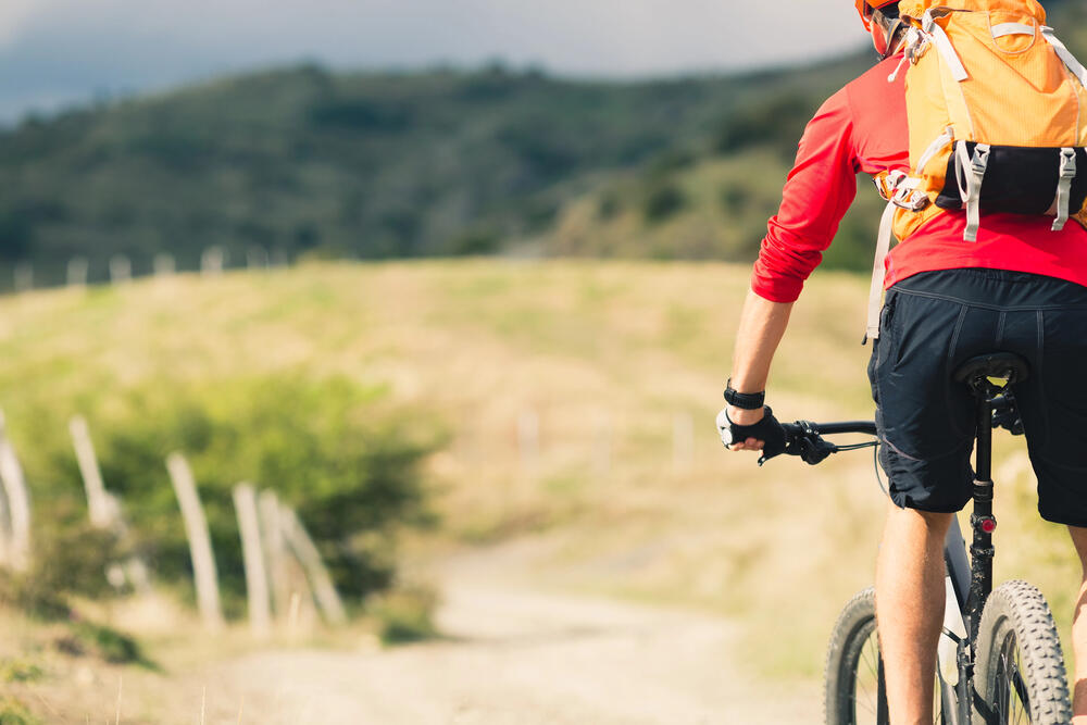 Cyclist riding along a rural dirt path through rolling hills, wearing a bright orange backpack and red shirt, capturing the immersive and slow-paced experience of exploring a landscape by bike.