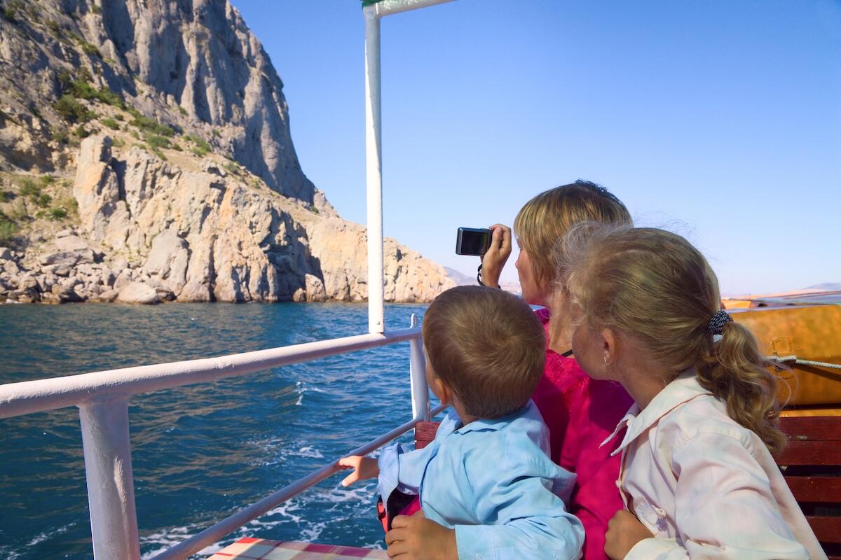 Three children enjoy a scenic boat ride, with one taking photos of rocky cliffs rising above the blue sea—highlighting a budget-friendly family adventure packed with exploration and memories.