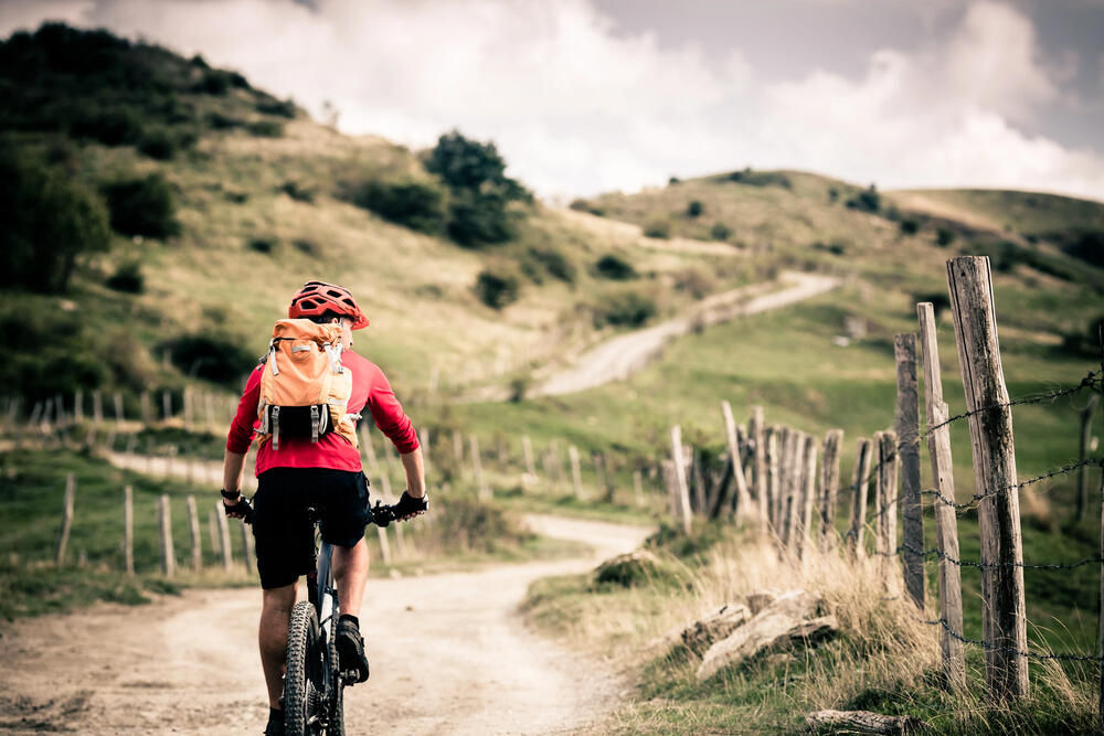 Cyclist with an orange backpack riding a mountain bike along a winding dirt path through scenic green hills, highlighting the slow, immersive experience of exploring remote landscapes by bike.