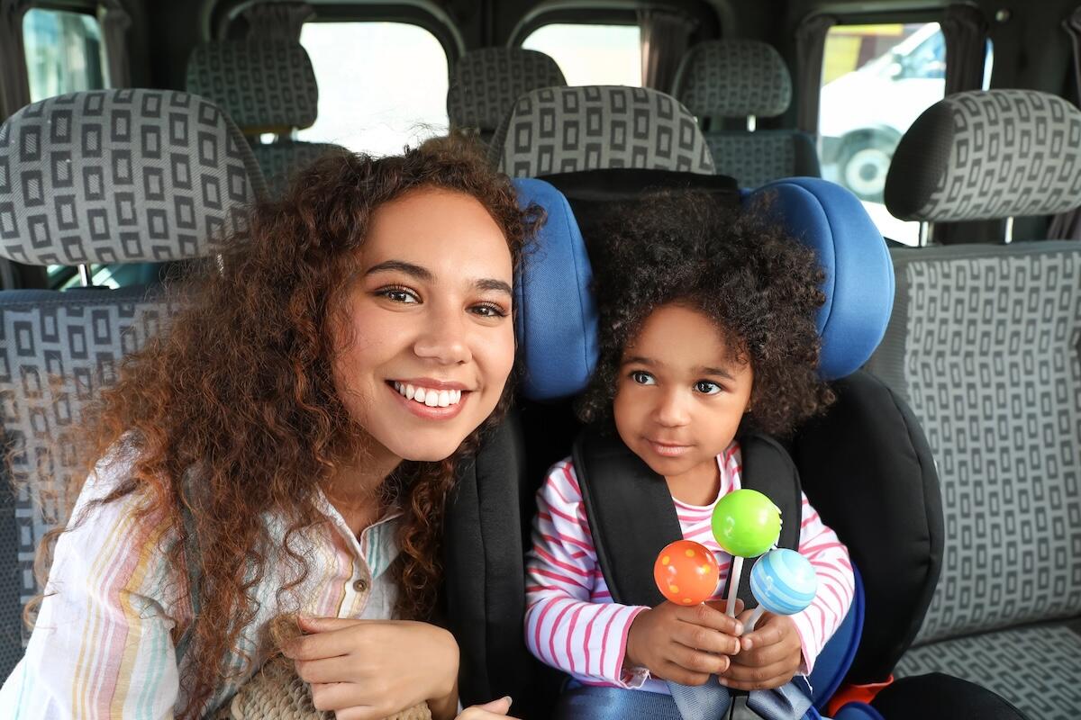 A smiling mother and her young daughter sitting in the backseat of an airport taxi. The child is safely strapped into a car seat and holding colourful toys, capturing a joyful moment during their solo mum travel adventure.