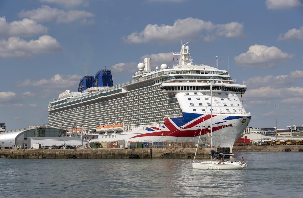 P&O Cruises' Britannia docked at the Port of Southampton, preparing to depart on a mini cruise, with a small sailboat passing in the foreground under a bright blue sky.